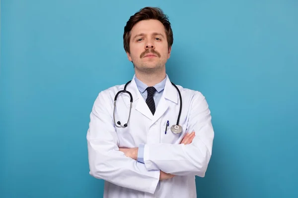 Молодой серьезный врач со стетоскопом позирует на синем фоне — стоковое фото
