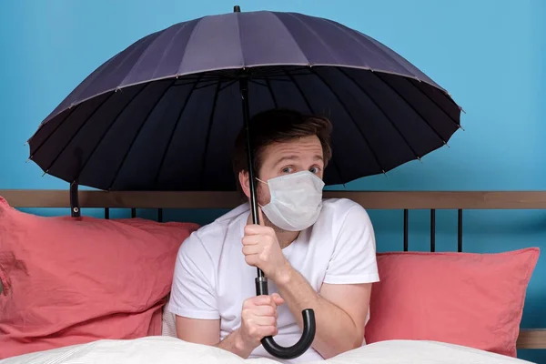 Чоловік у медичній масці тримає парасольку, сидячи на ліжку — стокове фото