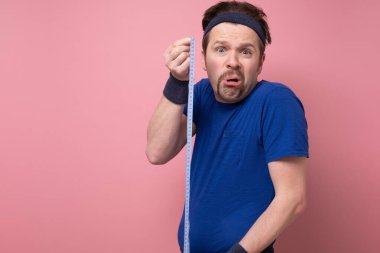 Kafası karışmış zayıf bir adam vücudunu santimetre santim ölçüyor. Trans sürecinin sonuçlarını görmediği için şaşırmış görünüyor. Stüdyo resmi pembe duvarda.