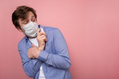 Coronavirus protection and disinfection or OCD concept. Caucasian man in medical mask spraying sanitizer spray over pink background clipart