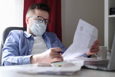 Tıp maskeli genç beyaz adam evde dizüstü bilgisayarla faturaları ve belgeleri karıştırıyor. İşadamı bürosunda evrak işleriyle uğraşıyor. Evde kalıp kendini karantinaya al..