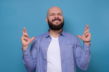 Pozitif sakallı adamın parmaklarını çaprazlamasına, mavi arka planda zevkle gözlerini kapatmasına sevindim. Mutlu erkek, rüyalarının gerçek olmasına sevinir. Olumlu yüz insani duygu.