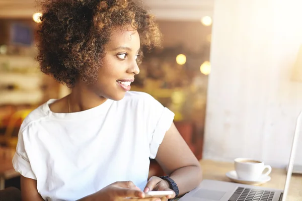 People, leisure and technology. Confident dark-skinned girl with Afro hairstyle wearing white top texting friends using smart phone, smiling, sitting at cafe table with cup of coffee and laptop