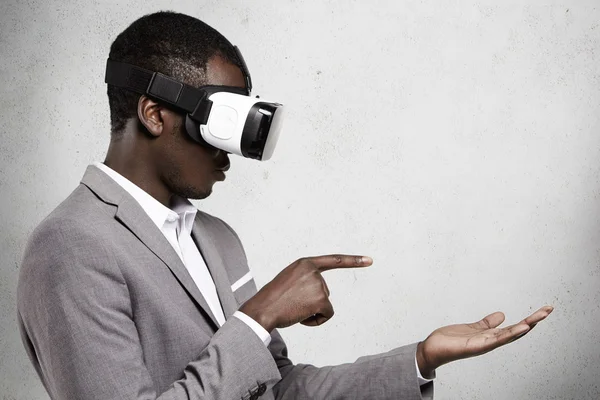 People, technology, entertainment and cyberspace concept. Young African businessman wearing suit and virtual reality headset, gestruring as if using some digital gadget while playing video game