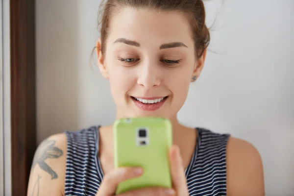 Close-up portrait of European girl playing games at her green smartphone. Young and charming woman holding her gadget with two hands, looking at display with slight smile. Indoors daylight shot.