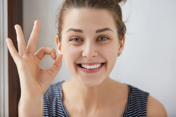 Young European brunette female showing OK-gesture with her fingers. Happy woman in striped top smiling with blue eyes. Her white-teeth mouth and happy face prove everything goes according to plan.