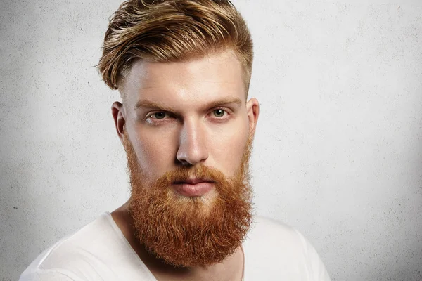 Portrait of young bearded dandy with hair brushed back in a trendy manner. Attractive Caucasian man in white T-shirt looking ahead, his charming green eyes sparkling. Indoors shot in white studio.