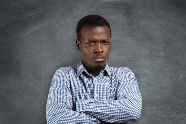 Headshot of angry serious African teacher with arms folded, dissatisfied with his misbehaving students, standing against blank chalkboard with copy space for your text or advertising content