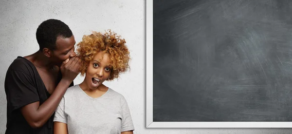 Handsome young African student in black t-shirt standing at blackboard, whispering something into ear of stylish girl, sharing latest gossip. Woman looking in shock and surprise with mouth wide open — Stock Photo