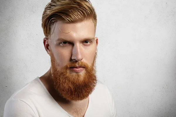 Portrait of fashionable young model in white t-shirt posing isolated against studio wall background. Bearded guy with stylish haircut dressed casually, looking serious and confident at camera