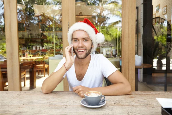 Young Caucasian man with happy attractive smile in red hat with white fur speaking on mobile phone with his friends while drinking coffee at cafe, spending Christmas holidays away in tropical country