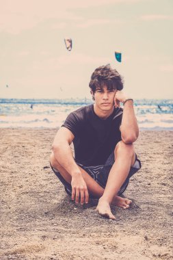 Tenerife plaj üzerinde iyi adam siyah t-shirt ile otur. Mavi okyanus ve dalgaları arkasında, sıcak vintage filtre. Kameraya bak.