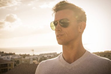 güzel genç adam gözlüklü aviator modeli ön bakmak hoşuna gidiyor. arka planda okyanus ve yüksek çatı üzerinden muhteşem manzaralı güzel altın gün batımı