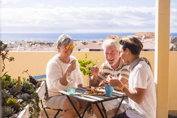 three people different ages eating and enjoying together the rooftop with sea view. Summer lifestyle with amazing panorama. Eat and drink food and beverage.