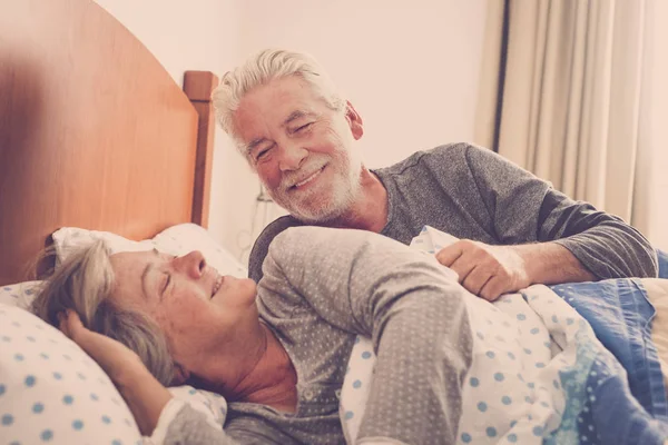 Couple of senior man and woman waking up and smiling with a hug while are in the bed at home. Vintage filter and light in the back. The man kiss the woman with love