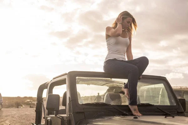 自由と オフロードの屋根の上に座って美しい金髪白人若い女性の休暇の概念は黒の車です バックライトと温かみのある色調の日光 — ストック写真