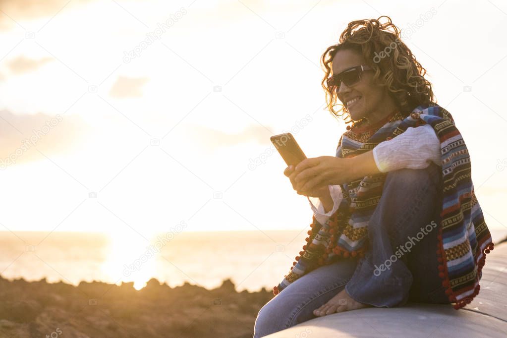 beautiful woman in silhouette during a golden amazing sunset on the ocean using a technology mobile smart phone sitting down on a long stone beach nice leisure in vacation writing friends
