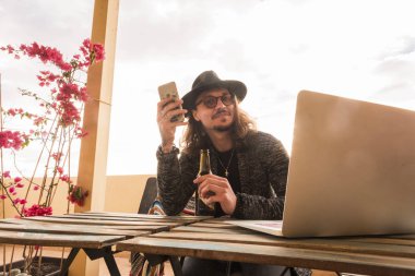 şapka ve gözlük trendy adamla arkadaşları ve insanlar ile iletişim kurmak için telefon kullanım sırasında bir bira iç. Laptop ücretsiz çalışmak için tablo açık terasta