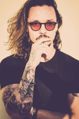 beautiful man with long black hair look at you thinking about it. Fashion model concept with vintage filter and warm tones. Black t-shirts and eyeglasses, trendy look and tattoos clipart
