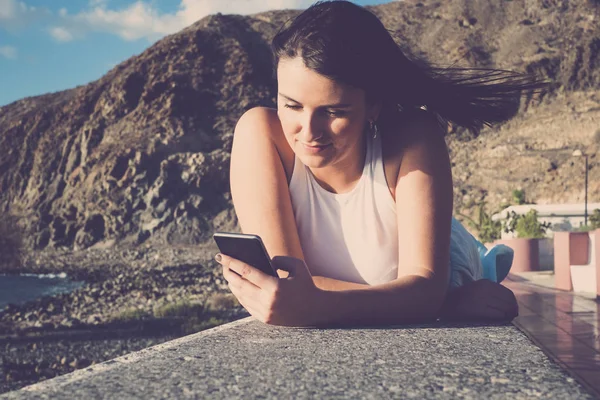 美丽的长发西班牙妇女使用手机发送信息在特内里费岛度假 躺在海边的墙上 阳光下的蓝天下 — 图库照片