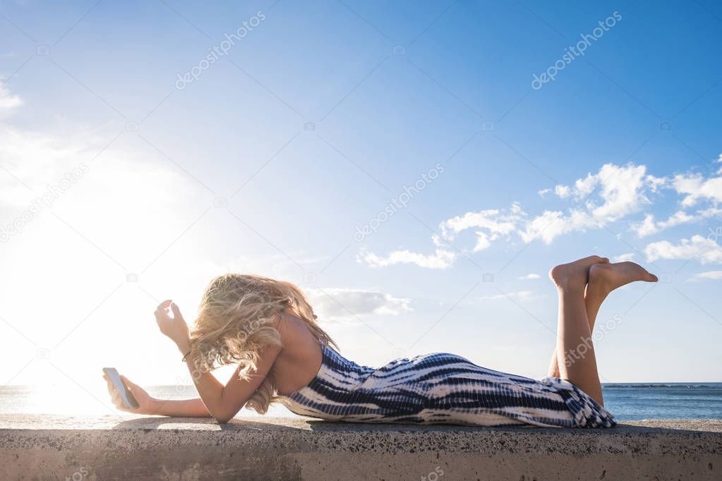 Beautiful long hair spanish woman use mobile phone to send message during a vacation in Tenerife. Lay down on a wall near the beach under a blue sky with sun.