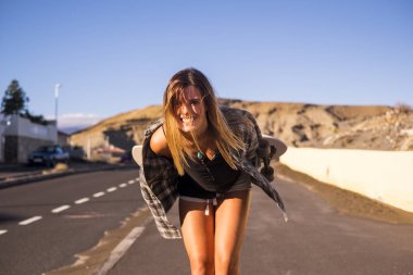 sarı uzun saçlı şirin model güzel gülümseme size kamera bir kaykay ile seyahat için uzun bir yolda yürüyor. dağlar gün tatil Tenerife arka plan ve güneş ışığı