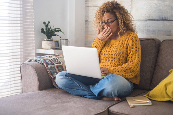Surprised adult woman at home sitting down on sofa with personal laptop computer 