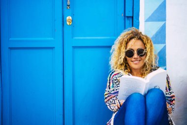 Mutlu, mutlu, kitap okuyan ve eğlenen bir kadının mavi portresi. Arka planda ev ve kapı olan yere otur. 