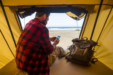 Man sit down inside a tent on wild free camping at the beach using modern connected cellular mobile phone 