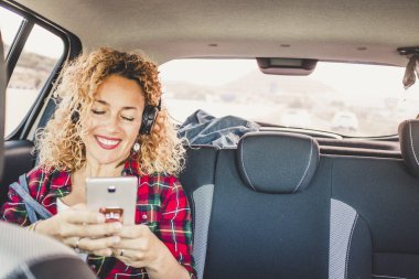 Neşeli, mutlu bir kadın modern bir arabanın arka koltuğunda oturup kulaklıklı müzik dinlemek için cep telefonu kullanıyor.