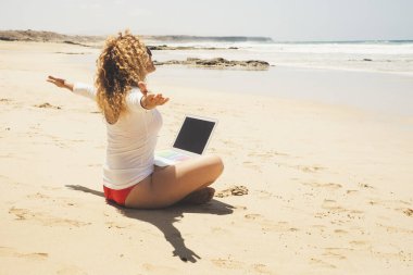 Mutlu ve özgür bir kadın sahilde dizüstü bilgisayarı bacaklarında oturuyor.