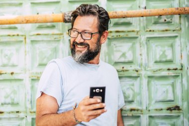 Yetişkin beyaz adam portresi cep telefonu cihazı kullanıyor ve arkasına bakıyor - arka planda yeşil eski kapı - insanlar ve açık hava teknolojisi