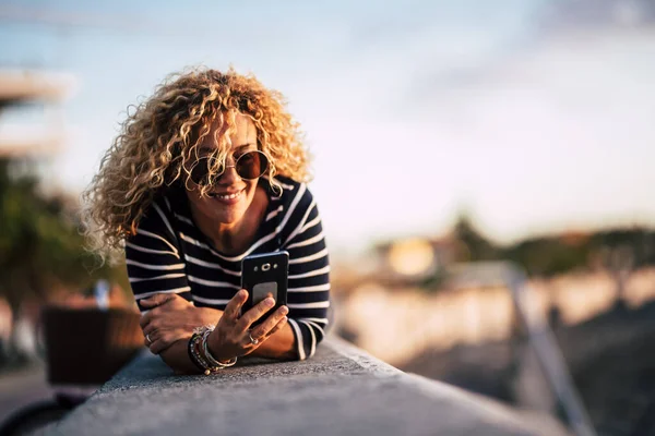 Beautiful caucasian blonde curly woman lay down outdoor and use cellular mobbile phone sending messages and reading internet connection news - people and technology