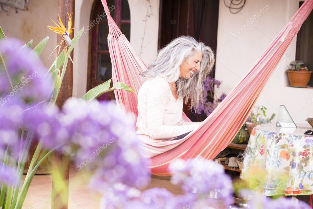 White hair diversity senior woman relax on an hammock in the garden at home reading a paper book and enjoying the quaity of leisure time alone