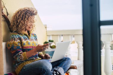 Güzel, beyaz, sarışın, kıvırcık, modern kadın evde telefon ve kişisel dizüstü bilgisayar kullanıyor. İnternet bağlantısı ve gezinme cihazı kavramı. Beleş ofis evi akıllı iş kavramı.