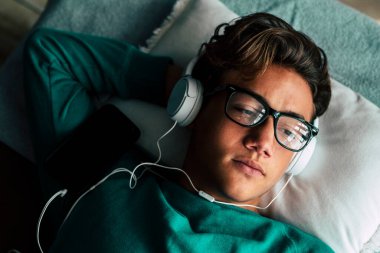 Sıkılmış yakışıklı, beyaz, genç bir erkek. Evde yatağa uzanıp kulaklıkla müzik dinliyor. 