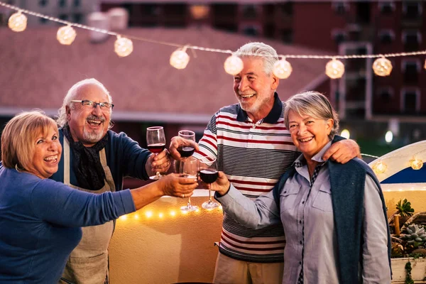 Happy group of old senior caucasian friends celebrate together by night clinking and toasting with red wine