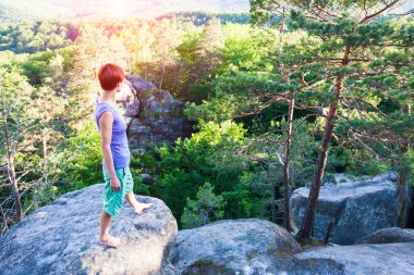 Uçurumun yakınındaki bir kaya üzerinde duruyor ve manzaraya görünüyor.