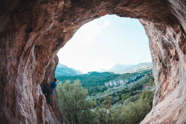 A man climbs the rock against of a mountain landscape. The athlete trains on a natural relief. Climbing in Turkey. Rock in the form of an arch. Cave. The climber overcomes a difficult route.