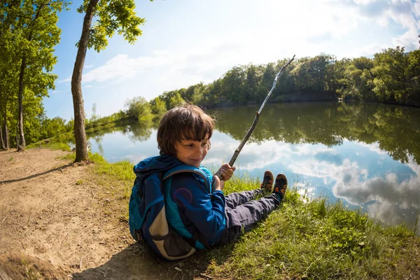 Un niño con una mochila se sienta en el lago con un palo de madera . — Foto de Stock
