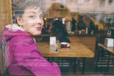 Gülümseyen bir kız bir kafede oturur ve pencereden dışarı bakar..