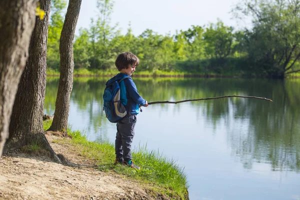 Un niño con una mochila se para en el lago con un palo de madera . — Foto de Stock