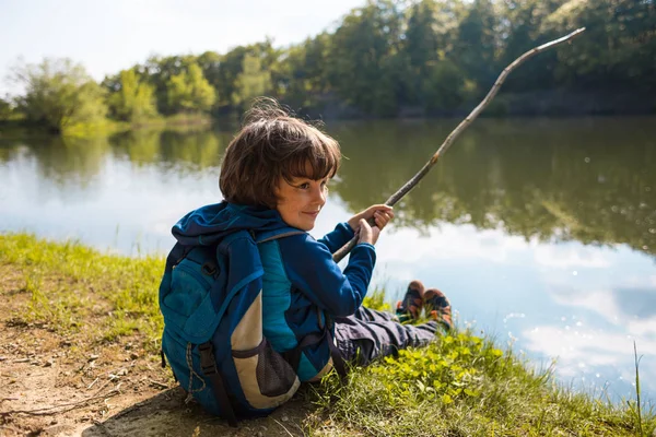 Un niño con una mochila se sienta en el lago con un palo de madera . — Foto de Stock