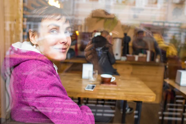 Una chica sonriente se sienta en una cafetería y mira por la ventana . — Foto de Stock