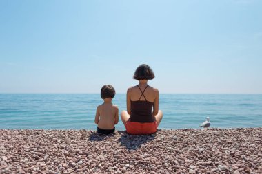 Annesiyle kumsalda oturup denize bakan bir çocuk..