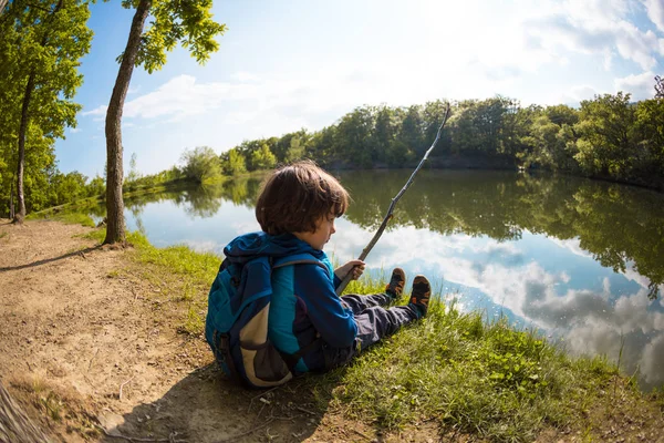 Chlapec s batohu sedí na jezeře s dřevěnou holí. — Stock fotografie