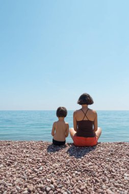 Annesiyle kumsalda oturup denize bakan bir çocuk..