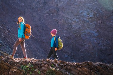 Bir kadın ve çocuğu dağ yolunda yürüyor. Sırt çantalı bir çocuk annesiyle seyahat eder. Çocuklarla yürüyüş. Çocuk ormanda yürür. Aktif yaz tatili. 