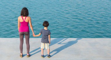 Annesiyle birlikte gezinti güvertesinde duran bir çocuk suya bakıyor. Şehir caddesinde oğluyla yürüyen bir kadın. Genç bir anne bebeğinin elini tutar..