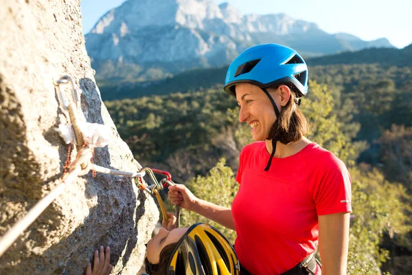 Mother teaches a child how to use safety equipment. A boy in a helmet goes through Via Ferrata. A woman instructs how to use a carabiner for belaying. Mountain tourism and mountaineering for children.